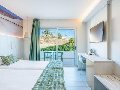 Camera d'hotel luminosa con due letti, decorazioni verdi e balcone con vista sulle palme.