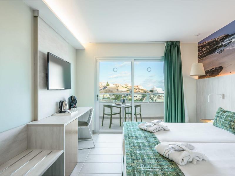 Camera d'hotel luminosa con letto matrimoniale, TV, scrivania e balcone con vista mare.