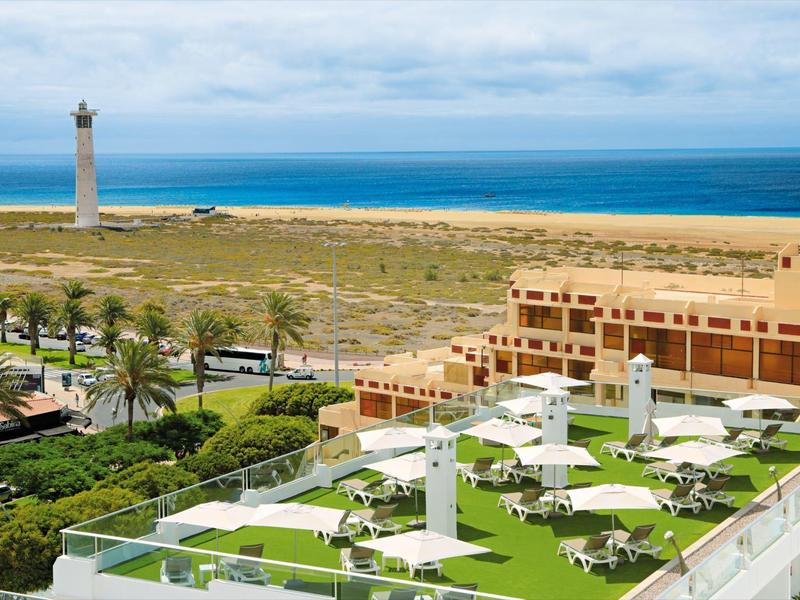 Vista di un hotel con prati verdi, ombrelloni e un faro sullo sfondo vicino al mare.