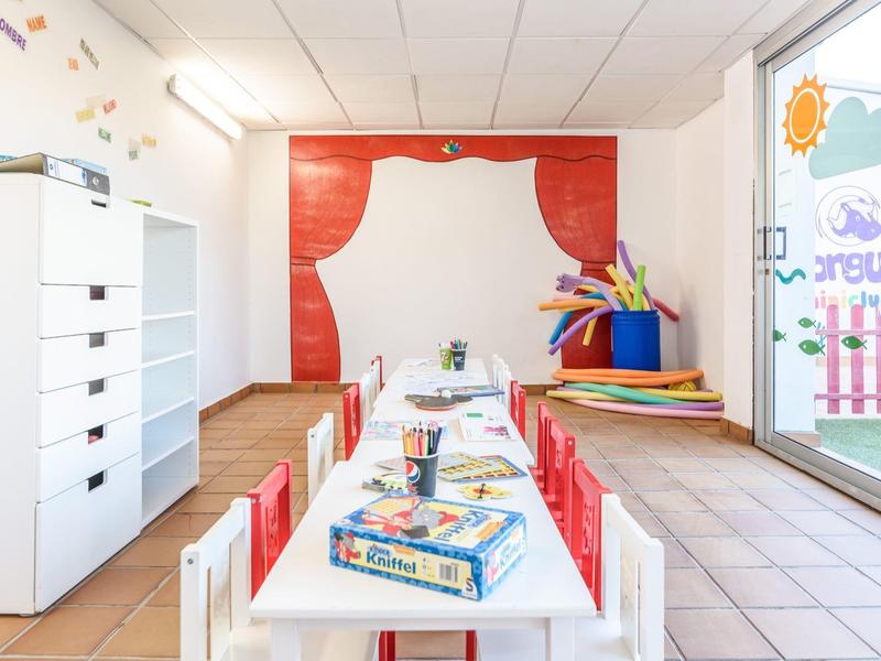 Area giochi per bambini con tavoli, sedie e giocattoli colorati in una stanza luminosa.