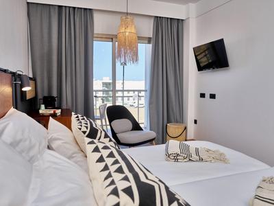 Helles Schlafzimmer mit Doppelbett, grauem Sessel, grauen Vorhängen und Fernseher an der Wand.