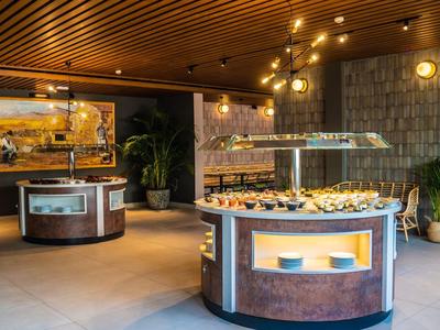 Restaurant mit warmem Licht, Holzdecke, Steinwand und runden Buffetstationen mit Speisen.