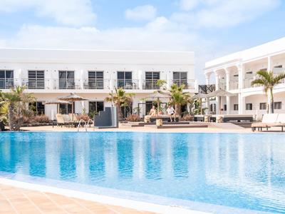 Großer blauer Swimmingpool vor weißem Hotel mit Palmen und Sonnenschirmen unter blauem Himmel.