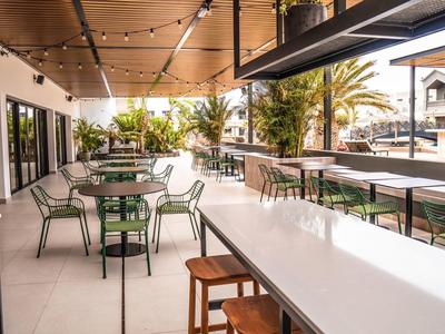 Moderne Außenbar mit weißen Tischen, grünen Stühlen und warmem Holzdekor unter Lichterkette.