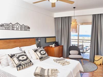 Helles Schlafzimmer mit großem Bett, gemusterten Kissen, Holzdeko und Balkon mit Meerblick.