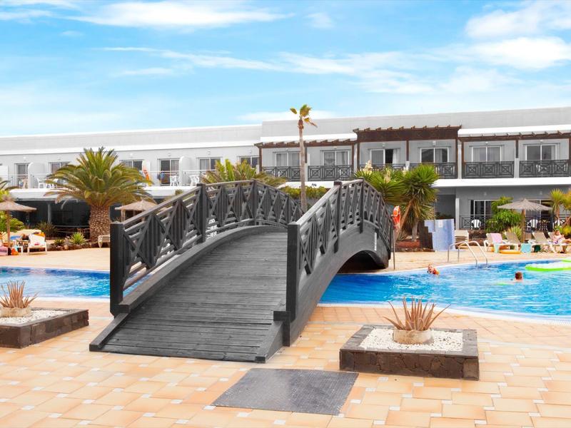 Zona piscina del resort con un piccolo ponte ad arco sull'acqua e edifici bianchi sullo sfondo.