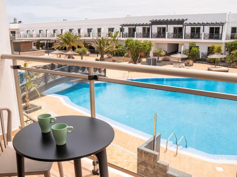 Balcone con tavolo e piante che si affaccia su una grande piscina all'aperto e un hotel moderno