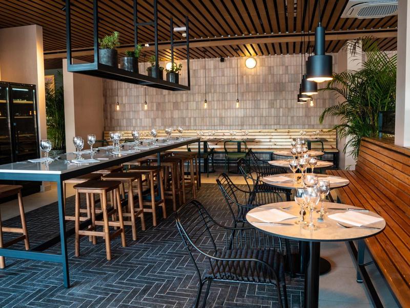 Interno di ristorante moderno con tavoli in legno, sedie e illuminazione soffusa contro un muro di pietra.