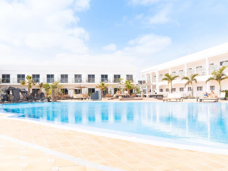 Ampia piscina di hotel luminosa con acqua limpida, circondata da lettini e edifici bianchi.