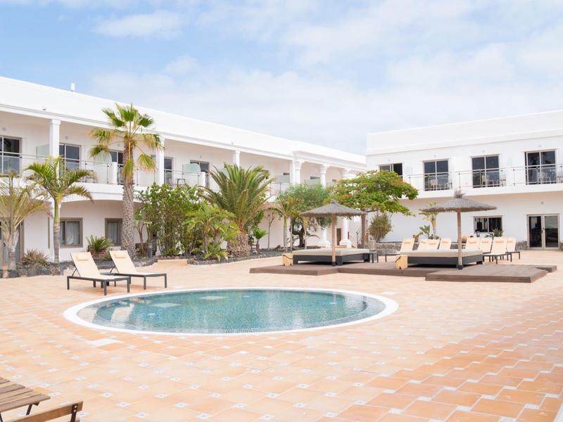 Cortile dell'hotel con piccola piscina rotonda, lettini, palme e edificio bianco su due piani.