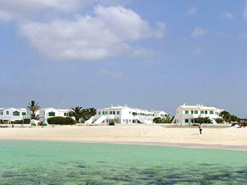 Strand mit weißem Sand, türkisfarbenem Wasser und weißen Gebäuden im Hintergrund unter blauem Himmel.