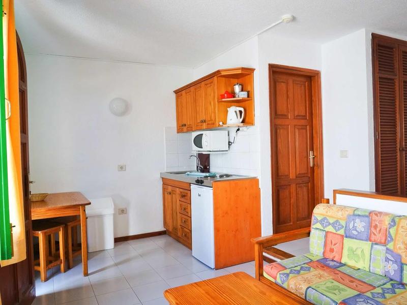 Kleines, helles Wohnzimmer mit Holz-Möbeln, bunter Couch, Tisch, Küchenzeile und braunen Türen.