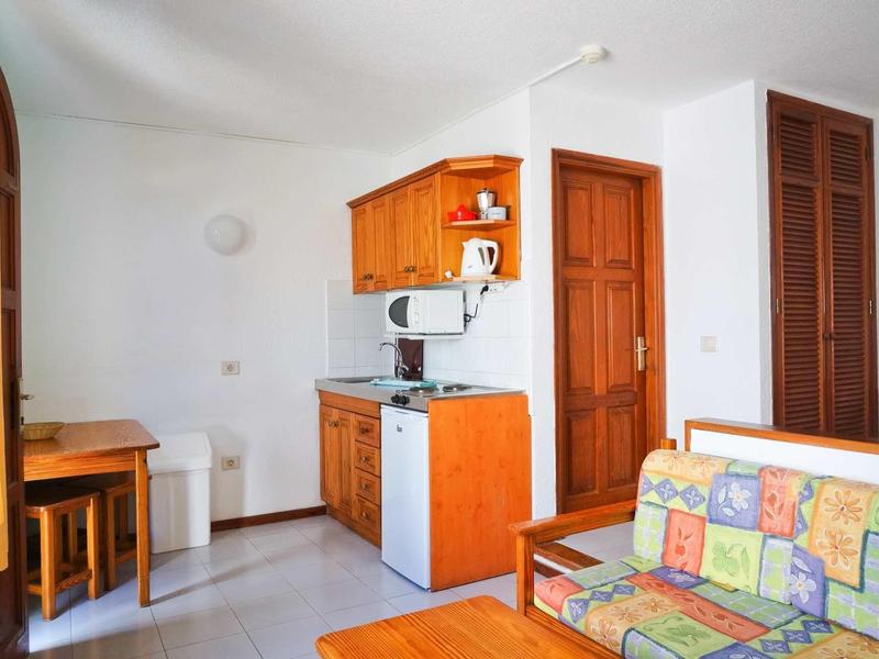 Kleiner Wohnraum mit Küchenzeile, Holzmöbeln und buntem Sofa vor weißen Wänden.