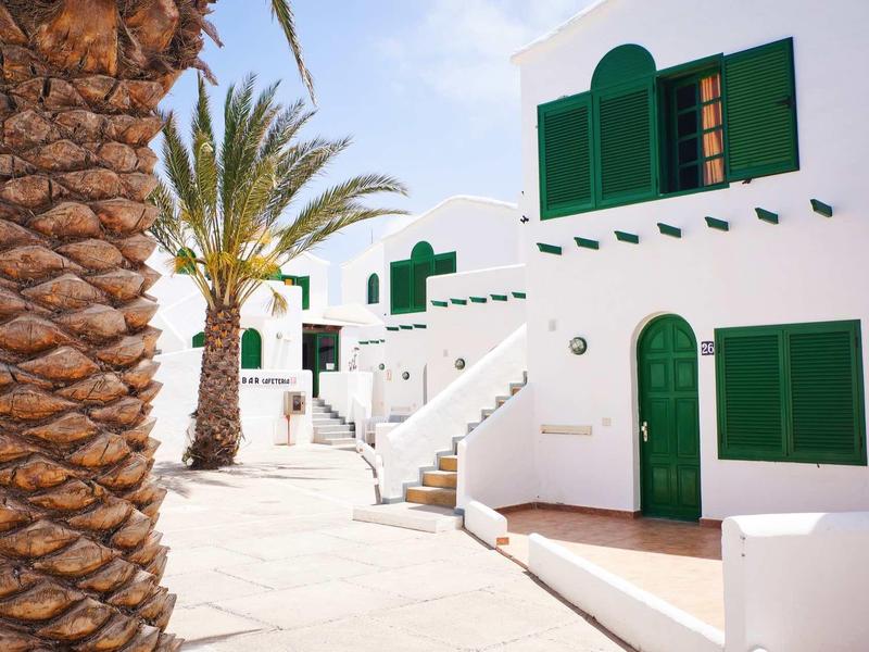 Weiße Häuser mit grünen Fenstern und Türen an einem sonnigen Tag, Palmen am Wegesrand.