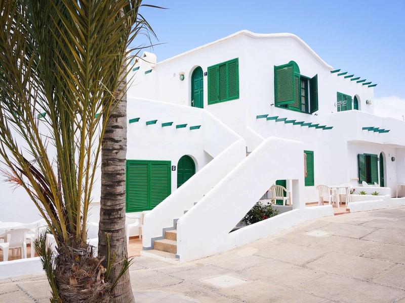 Weißes Haus mit grünen Fensterläden und Tür, Palmen und heller Terrasse darunter.