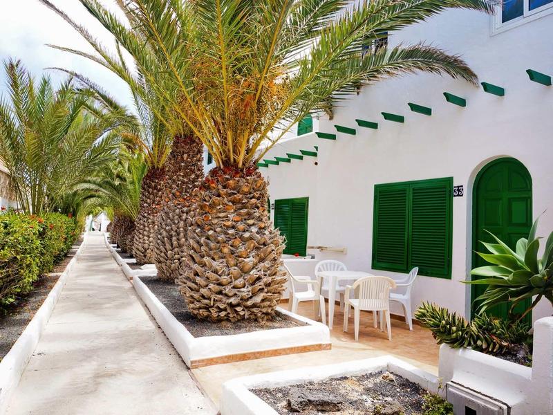 Weißes Haus mit grünen Fenstern, Palmen und weißen Außenmöbeln an sonnigem Tag.