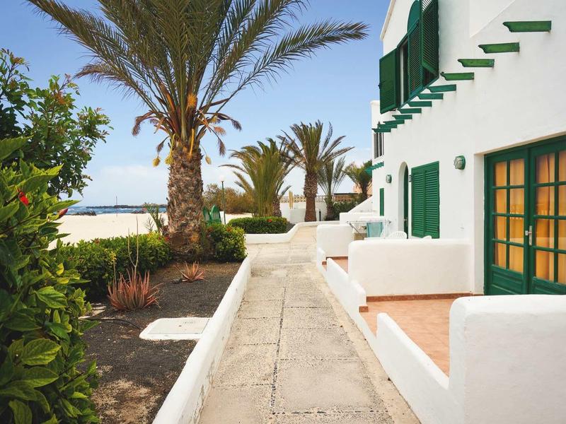 Schmaler Weg entlang weißer Häuser mit grünen Fensterläden, Palmen und Pflanzen bei klarem Himmel.