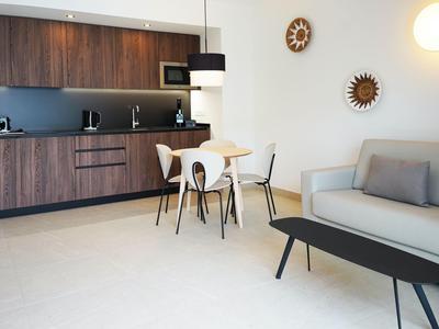 Modern hotel living area with kitchenette, dining table, sofa, and decorative wall lighting.