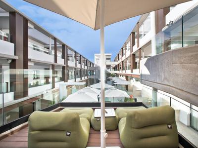 Area hotel moderna con piscina, sdraio e parapetti in vetro sotto un cielo azzurro.