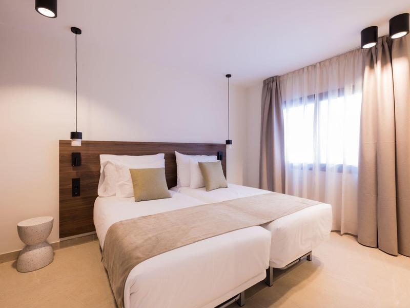 Camera d'hotel luminosa con letto matrimoniale, panca e tende grandi alla finestra.