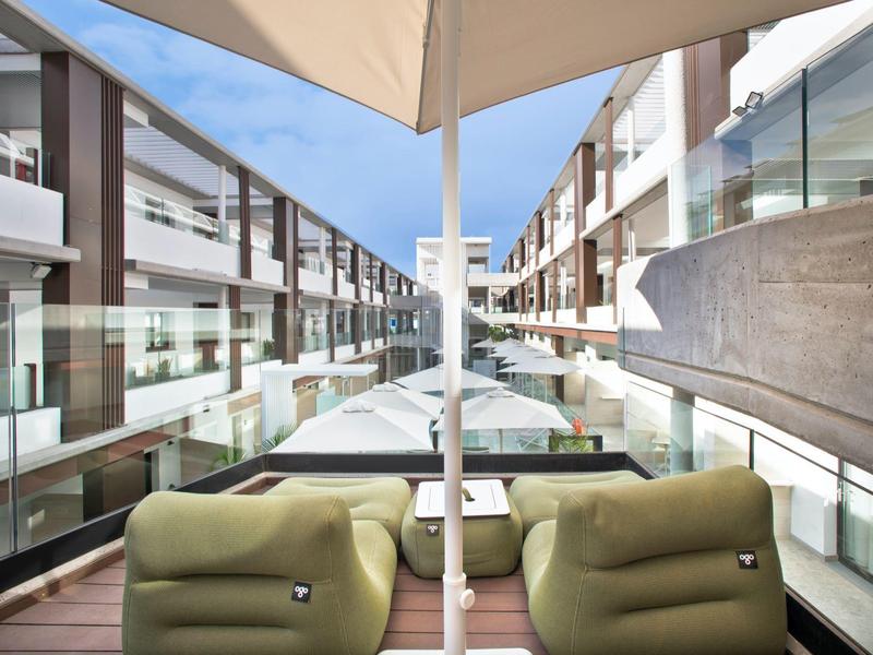 Area hotel moderna con piscina, sdraio e parapetti in vetro sotto un cielo azzurro.