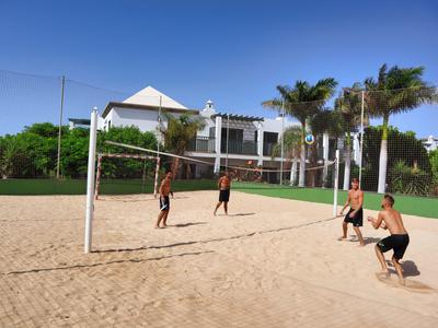Vier Personen spielen Beachvolleyball auf einem Sandplatz vor einem weißen Gebäude mit Palmen.