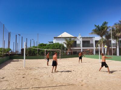 Vier Männer spielen Beachvolleyball auf einem Sandfeld vor einem Hotel unter klarem Himmel.