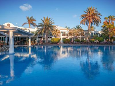 Großer blauer Pool vor einem Hotel mit Palmen und klarem Himmel.