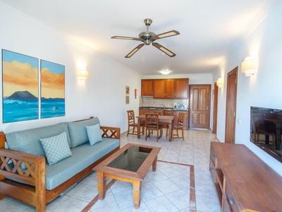 Soggiorno luminoso con divano in legno, ventilatore a soffitto e cucina aperta sullo sfondo.