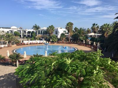 Rundes Pool mit Wasserfontänen umgeben von Palmen und weißen Liegestühlen in einem Hotelresort.