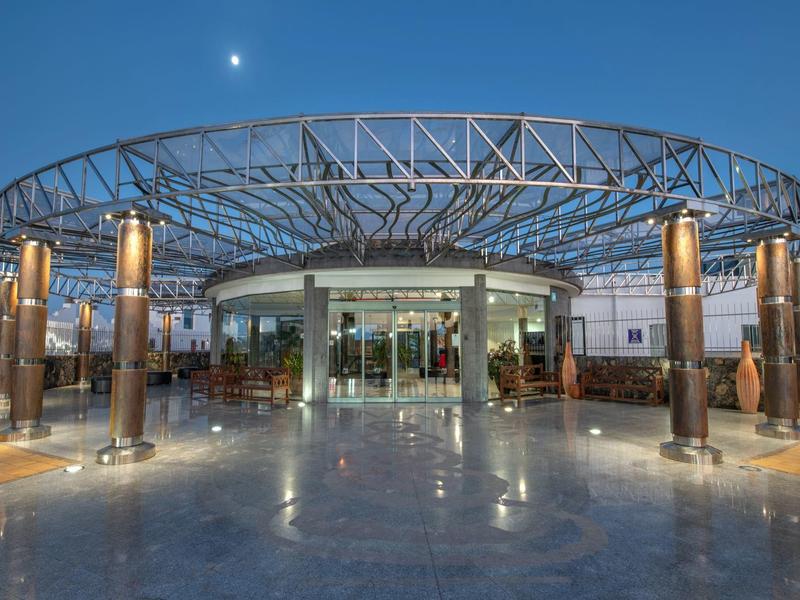 Vista serale dell'ingresso di un hotel moderno con facciata in vetro illuminata e struttura in acciaio circolare.