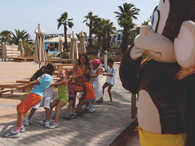 Bambini giocano su un'area lastricata mentre una persona in costume da animale osserva.