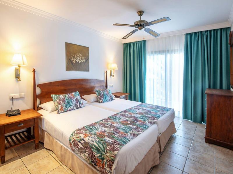 Camera d'hotel luminosa con due letti singoli, testiere in legno, ventilatore a soffitto e grandi tende verdi.