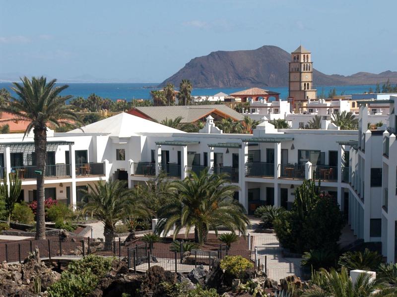 Hotel con facciata bianca, palme e montagne sullo sfondo vicino al mare.