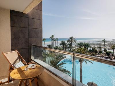 Balkon s dřevěnou židlí a stolem, v pozadí moře a bazén, jasná obloha.