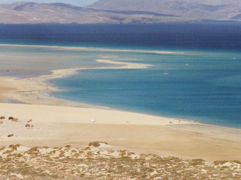 Zandstrand met helder blauw water en verre bergen onder een heldere lucht.