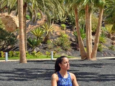 Femme médite détendue sur tapis de yoga en plein air devant un paysage tropical avec des palmiers.