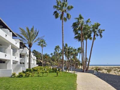 Complejo hotelero moderno con edificios blancos, palmeras y camino junto a la playa bajo cielo azul claro