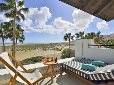 Balkon mit Liegestuhl und Liege, Blick auf Palmen, Sand und Meer unter blauem Himmel.