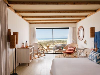 Helles Hotelzimmer mit Holzdecke, Doppelbett, Sitzbereich und Meerblick durch große Fenster.