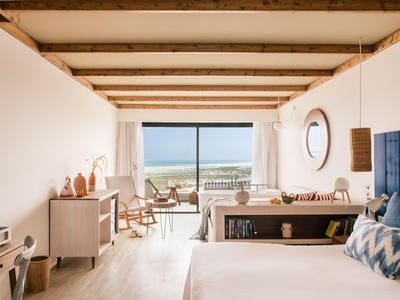 Camera d'hotel luminosa con soffitto con travi in legno, letto matrimoniale, area salotto e vista mare attraverso grandi finestre.