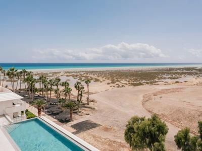 Blick auf einen Pool und Palmen am Hotel mit Blick auf einen weiten Strand und Meer unter klarem Himmel.