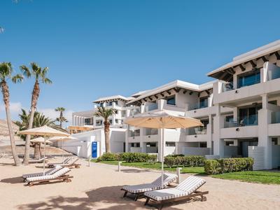 Hotel mit weißen Häusern und Liegen am sonnigen Strand unter blauem Himmel.