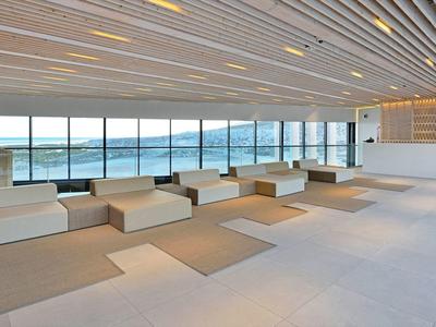 Área de espera moderna y luminosa con bancos blancos y grandes ventanas con vista al mar.