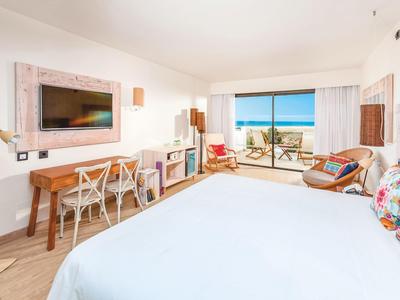 Habitación de hotel luminosa con vista al mar, cama grande, escritorio y balcón con asientos.