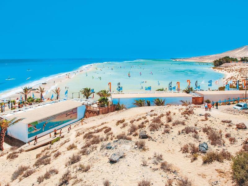 Panorama de una playa de arena concurrida con sombrillas y mar azul bajo un cielo despejado.