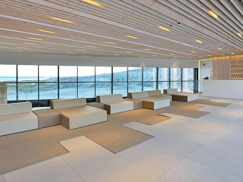 Área de espera moderna y luminosa con bancos blancos y grandes ventanas con vista al mar.