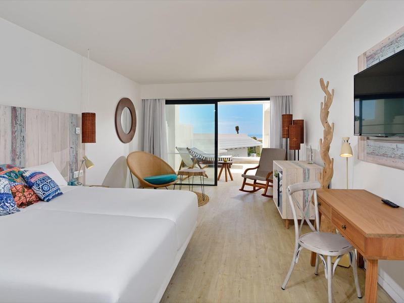 Habitación de hotel moderna con cama grande, escritorio, balcón y vista al mar durante el día.