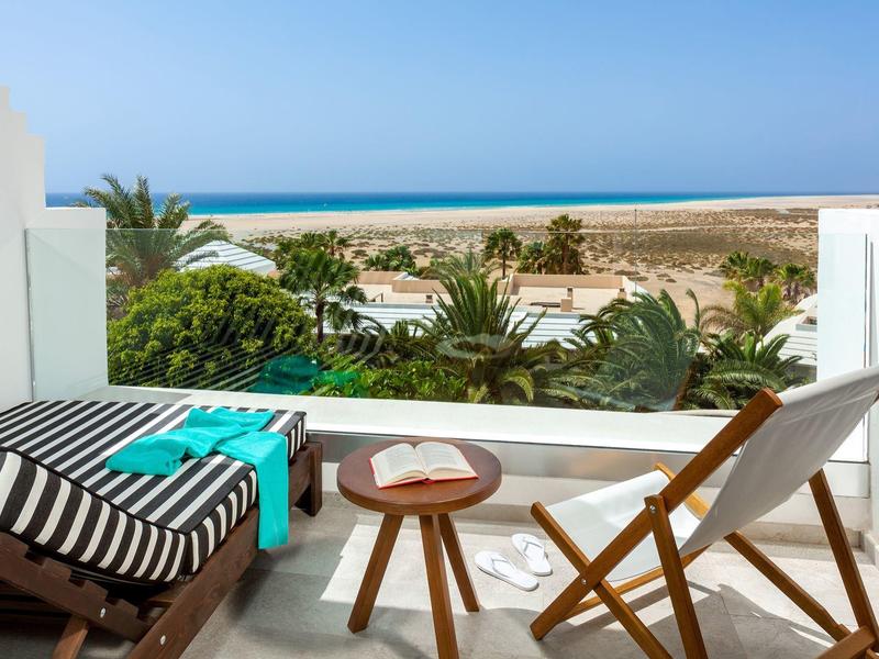 Balcón con sillas y mesa con vista a palmeras y el mar en un día soleado.