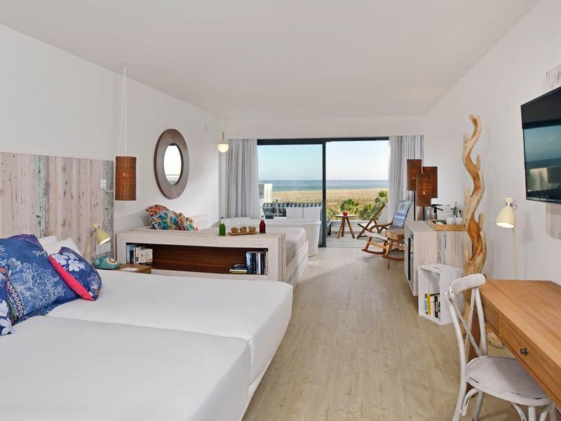 Suite de hotel luminosa con cama doble, muebles de madera y vista al mar a través de grandes ventanas.